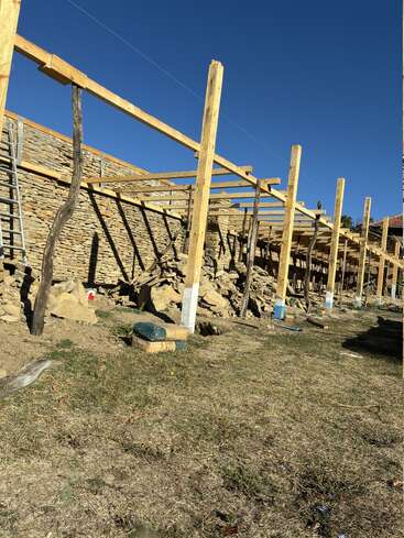 Dieses Bild zeigt eine Baustelle mit Holzbalken und Pfosten, die ein Gerüst bilden, vor einer Steinmauer. Das Gelände ist grasbewachsen und der Himmel ist klar und blau.