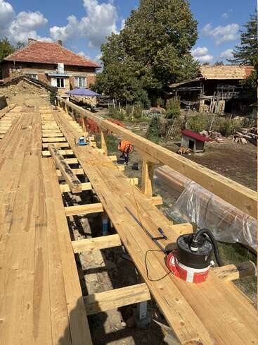 In einer ländlichen Umgebung wird eine Holzkonstruktion gebaut. Im Hintergrund sind Werkzeuge, ein altes Haus, Bäume und ein klarer blauer Himmel zu sehen. Einige Ziegelsteine und Geräte sind zu sehen.