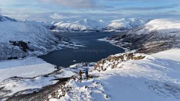 Two people hike along a snowy mountain ridge overlooking a stunning fjord; surrounding peaks and valleys are beautifully covered in snow under a bright, clear sky.