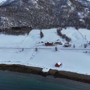 A snowy landscape with a mountain background, red houses, leafless trees, a road, a dock, and calm shoreline, creating a peaceful and wintery rural scene.
