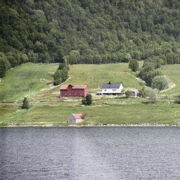 The image depicts a serene rural landscape featuring a cluster of houses on a hillside, overlooking a tranquil body of water and a lush forest in the background.