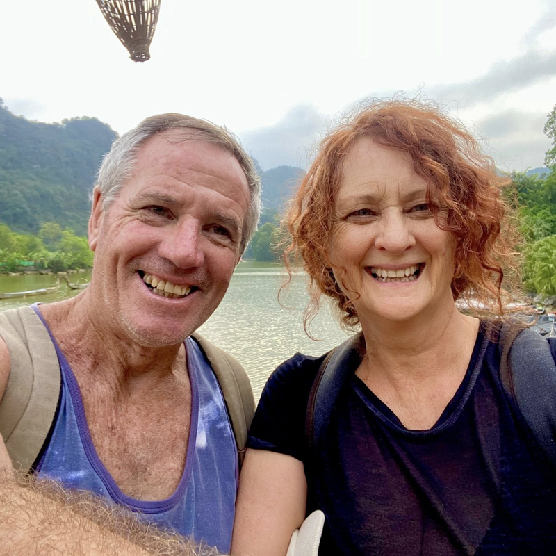 A smiling couple takes a selfie outdoors, standing by a lake with lush green trees and misty mountains in the background, both wearing backpacks and casual clothing.