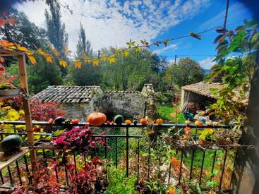 The image depicts a serene garden scene with a black metal fence, vibrant plants, and pumpkins, set against a backdrop of a stone house and a blue sky with white clouds.