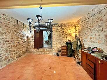 Cette pièce se caractérise par ses murs en pierre rustiques, son sol en terre cuite et un lustre original. On y trouve des commodes, des vêtements, divers objets et une porte ouverte.