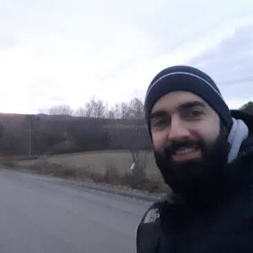 The image shows a man in a beanie and jacket standing on the side of a road, with a field and trees in the background on an overcast day.