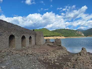 Esta imagen muestra antiguas ruinas de piedra junto a un lago sereno, rodeado de verdes colinas bajo un cielo azul brillante con nubes dispersas y esponjosas. Tranquilo e histórico.