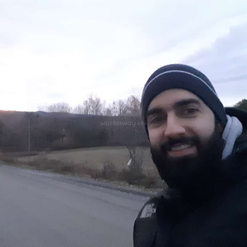 La imagen muestra a un hombre con gorro y chaqueta de pie al borde de una carretera, con un campo y árboles de fondo en un día nublado.
