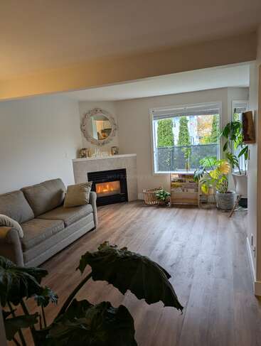 Ein gemütliches Wohnzimmer mit beigem Sofa, Holzboden, Kamin, großem Fenster, Zimmerpflanzen, Spiegel und minimalistischer Einrichtung schafft eine helle und einladende Atmosphäre.