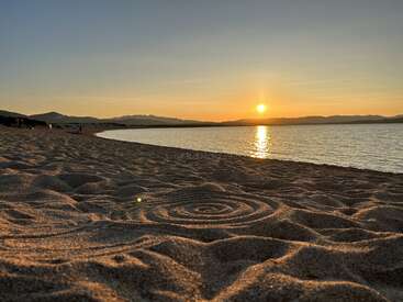 A imagem retrata uma cena serena de praia ao pôr do sol, com uma costa arenosa com padrões intrincados, um corpo de água tranquilo e um céu de tirar o fôlego com o sol se pondo sobre montanhas distantes.