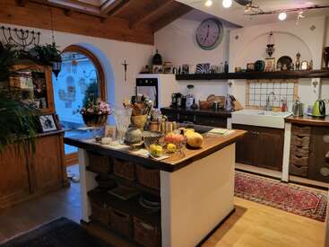 This cozy kitchen features wooden accents, a central island with fruit, baskets, and utensils, open shelves, a farmhouse sink, decorative items, and a large arched window.