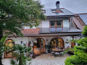 A charming house with solar panels on the roof, a tiled patio, outdoor seating, lush greenery, arched windows, and warm, inviting decor visible inside.