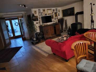 Cozy living room with a red sofa, wooden chairs, and a wall-mounted TV. Decor includes many framed pictures, indoor plants, rugs, and a fireplace.