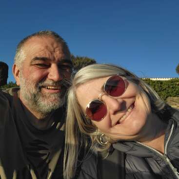 A smiling couple takes a cheerful selfie outdoors on a sunny day. The woman wears sunglasses, and both appear happy, relaxed, and enjoying each other's company.
