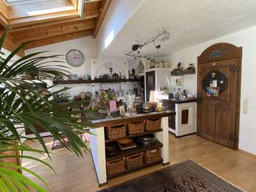 This cozy kitchen features wooden beams, a skylight, rustic decor, a wooden island with baskets, plants, a vintage clock, and a unique arched wooden door.