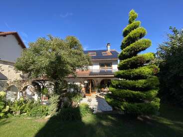 A charming house with arched patio, surrounded by lush green plants and unique spiral-shaped tree, under a bright blue sky on a sunny day.