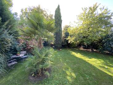 A lush, green garden with various trees, a palm in the foreground, shaded grass, and a cozy bench-table set in the left corner. Peaceful atmosphere.