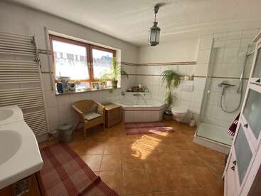 This spacious, sunlit bathroom features a corner bathtub, glass shower, double sinks, wicker chair, plants, warm tiles, red rugs, large window, and modern decor.