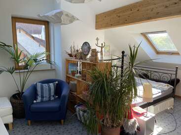 A cozy attic room with a blue armchair, bed, wooden bookshelf, several potted plants, decorative items, and natural light from two large windows. Inviting atmosphere.