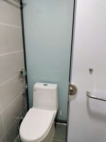 Esta imagen muestra un cuarto de baño moderno y compacto con un inodoro blanco, un bidé de mano, paredes de azulejos grises, mampara de cristal esmerilado y una puerta parcialmente abierta.