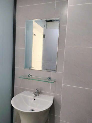 Esta imagen muestra un cuarto de baño moderno con azulejos de color gris claro, un espejo rectangular, una repisa de cristal y un lavabo blanco con grifo cromado.