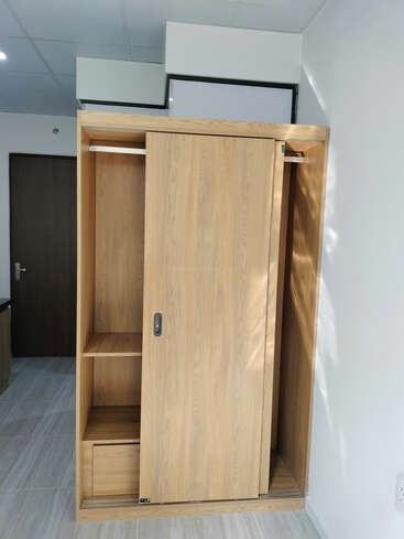 Esta imagen muestra un moderno armario de madera con una puerta corredera, estantes y una barra para colgar. Está colocado contra una pared blanca en una habitación luminosa.