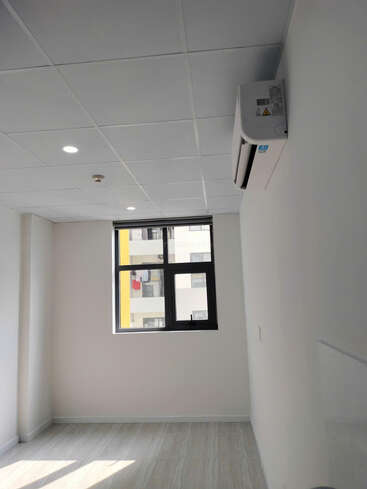 Esta imagen muestra una habitación pequeña, vacía e iluminada por el sol, con paredes blancas, suelo de baldosas, una ventana, un split de aire acondicionado y un techo abuhardillado con luces.