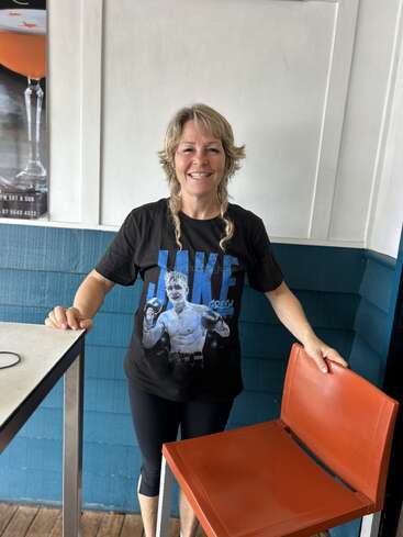 A smiling woman with blonde hair stands indoors, wearing a black T-shirt featuring a boxer. She holds an orange chair and stands beside a table.