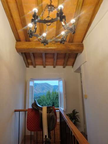 A cozy hallway with a wooden ceiling and chandelier, horse saddle on a railing, open window revealing lush green hills, and a potted plant in the corner.