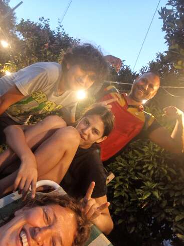 Four people pose outdoors at dusk, smiling and making playful gestures. Warm lights decorate the background, creating a cheerful, relaxed atmosphere among friends or family.
