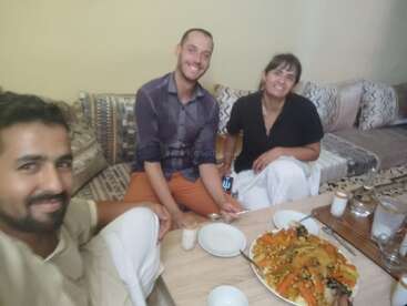 Three people are sitting together on a couch, smiling, and enjoying a meal. A large plate of colorful food and several drinks are on the table.
