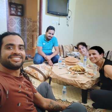 Quatre personnes sont assises sur des coussins de sol autour d'une table basse, souriant et dégustant un repas composé de pain, de frites, de salades et de bouteilles d'eau dans une pièce confortable.