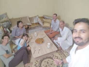 Un groupe de six personnes est assis autour d'une table basse, partageant nourriture et boissons, souriant joyeusement dans un salon confortable, chaleureusement décoré de coussins.