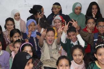 A group of joyful children, dressed in colorful traditional clothes, smile and make peace signs, creating a lively, cheerful atmosphere in a classroom setting.
