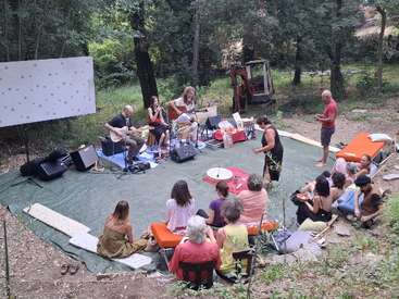 A group of people enjoy an outdoor music performance in a forest clearing. Musicians play guitars while a small audience sits on mats and cushions.