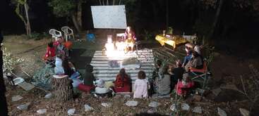 A group of people sit outdoors at night, facing a person illuminated on a small stage. The setting is cozy, surrounded by trees, rugs, and chairs.
