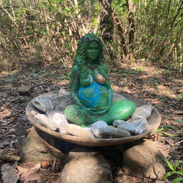 L'image représente une scène de forêt sereine avec une statue de femme enceinte, entourée de rochers et de feuilles, à l'expression sereine.