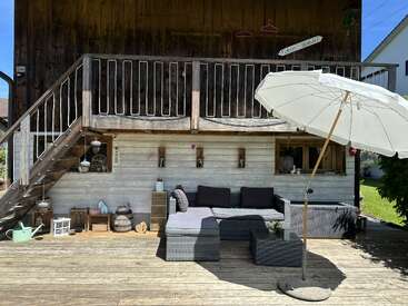 Eine gemütliche Holzterrasse mit Gartenmöbeln, einem Sonnenschirm, rustikaler Dekoration, Treppen und einem Balkon. Das Sonnenlicht erhellt den Bereich und schafft eine entspannte, einladende Atmosphäre.