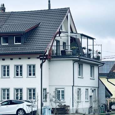 Ein modernes weißes Haus mit einem grauen Ziegeldach, mehreren Fenstern und einem oberen Balkon voller Pflanzen. Ein weißes Auto ist draußen geparkt. Bedeckter Himmel.