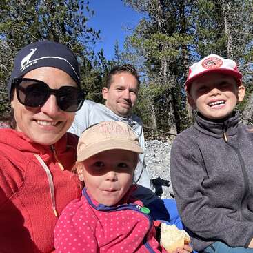 A smiling family of four enjoys a sunny day outdoors, surrounded by trees. The children look happy, wearing hats, while the parents wear sunglasses and casual attire.