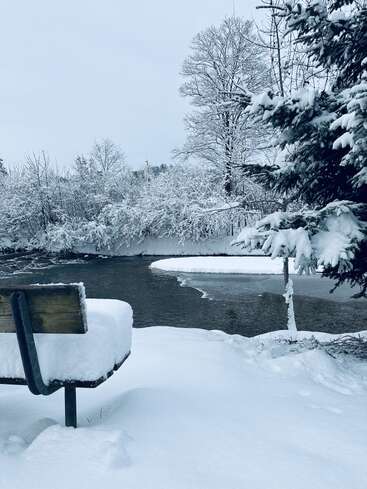 Eine schneebedeckte Bank mit Blick auf einen ruhigen, teilweise gefrorenen Fluss. Weiß bedeckte Bäume umgeben die Szene und schaffen eine friedliche Winterlandschaft unter einem bewölkten Himmel.