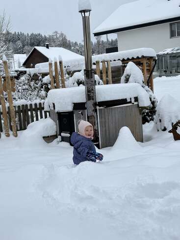 Ein kleines Kind in einem lila Mantel und einer rosa Mütze spielt fröhlich im tiefen Schnee in einem Hinterhof, umgeben von schneebedeckten Dächern und Bäumen in der Nähe.