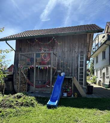 Ein rustikaler Holzschuppen ist mit bunten Wimpeln geschmückt. An einem sonnigen Tag gibt's da 'ne Hängematte, 'ne Kletterwand, 'ne blaue Rutsche, 'nen Holzstapel, 'ne Leiter und Kinderspielzeug.