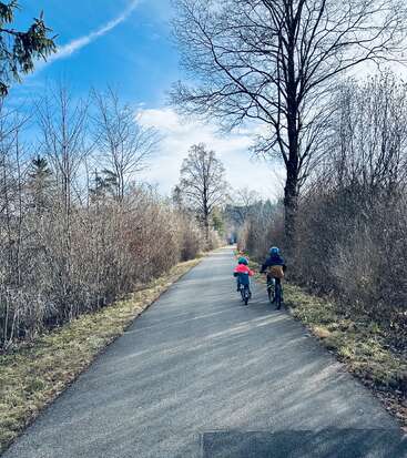 Zwei Kinder fahren mit ihren Fahrrädern einen gepflasterten Weg entlang, umgeben von kahlen Bäumen und Büschen unter strahlend blauem Himmel, und genießen gemeinsam ein friedliches, malerisches Abenteuer im Freien.