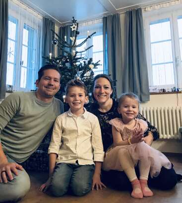 Eine lächelnde vierköpfige Familie steht drinnen vor einem geschmückten Weihnachtsbaum und genießt gemeinsam einen festlichen Moment. Es herrscht eine warme, fröhliche Atmosphäre mit natürlichem Licht.