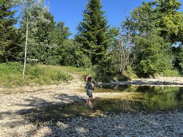 Ein Kind läuft barfuß an einem sonnigen Tag durch einen flachen, felsigen Bach. Üppig grüne Bäume umgeben die ruhige Gegend, und eine andere Person steht in der Nähe.