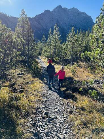 Zwei Kinder laufen Hand in Hand auf einem steinigen Waldweg, umgeben von Kiefern, unter einem strahlend blauen Himmel, mit majestätischen Bergen im Hintergrund.