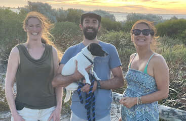 Trois personnes et un petit chien posent joyeusement en plein air au coucher du soleil, avec de la verdure et la mer en arrière-plan. Ils sourient et semblent détendus.