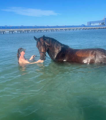 Une femme se tient debout dans l'eau claire jusqu'à la taille, tenant doucement un cheval brun. Le ciel est bleu et une longue jetée bleue s'étend à l'arrière-plan.