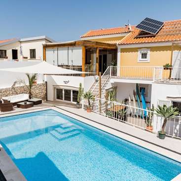 The image depicts a serene backyard scene featuring a rectangular swimming pool, a white patio umbrella, and a two-story house with a balcony and solar panels on the roof.