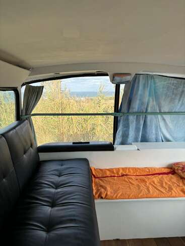 This image shows the cozy interior of a camper van, featuring a black leather couch, orange blanket, large windows, curtains, and an outdoor view of tall grass.